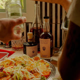 Flamingo Estate Balsamic Vinegar joins the Persimmon Vinegar at a citrus salad table, a hand reaching past a striped chair in warm light.