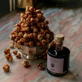 A Flamingo Estate Barrel Aged Balsamic Vinegar bottle stands on marble beside a silver footed bowl piled high with caramel-glazed popcorn.