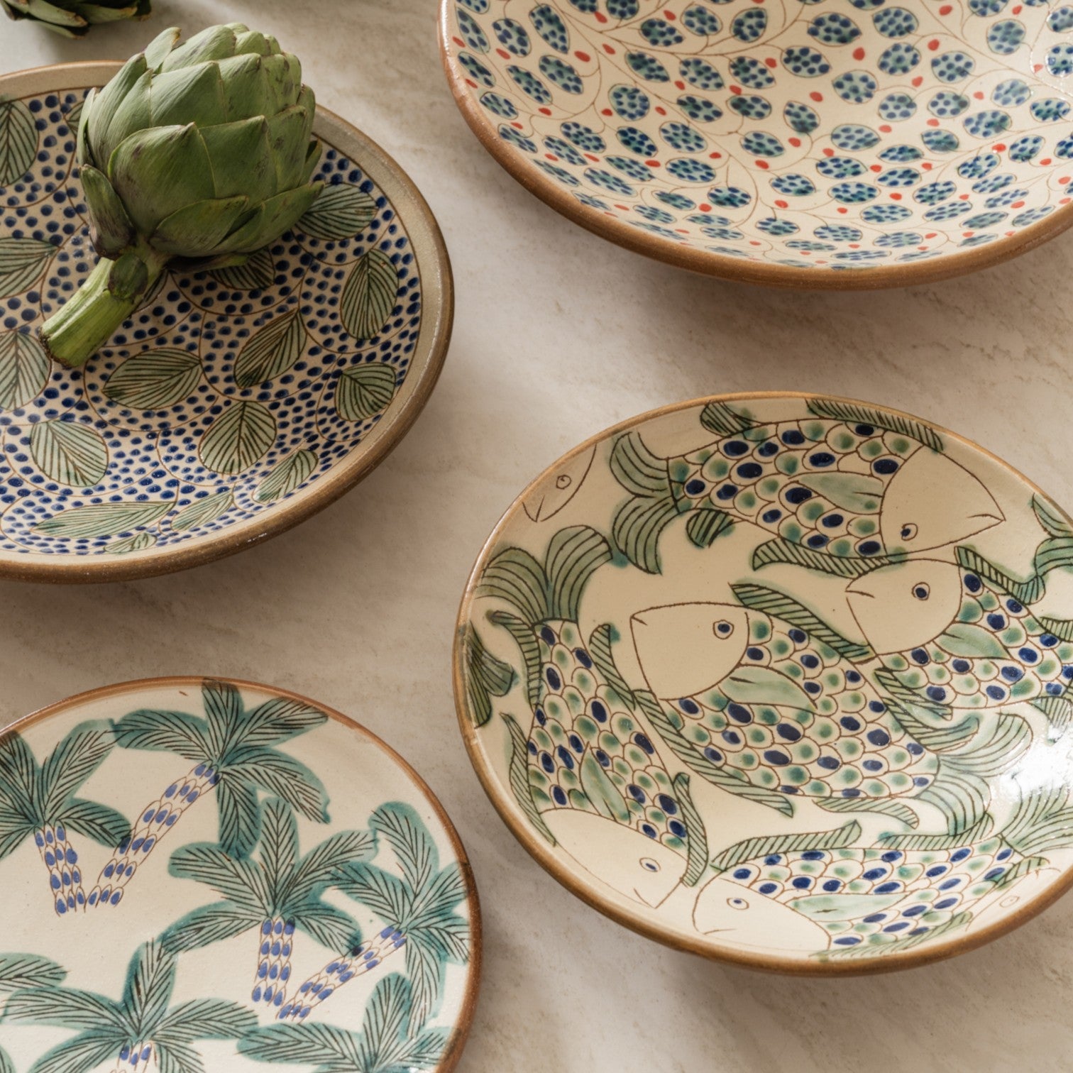 Calimia Home handcrafted ceramic plates featuring hand-painted palm, fish, and floral motifs on a marble surface.