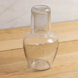 Be Home clear glass bedside carafe with a textured pebble finish and matching nesting tumbler on a wooden surface.