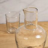 Be Home clear glass bedside carafe and matching tumbler with a textured pebble-like finish on a wooden surface.