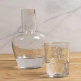 Be Home clear glass bedside carafe and tumbler set with a textured, pebble-like finish on a wooden surface.