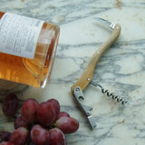 Be Home olivewood double-hinged corkscrew with stainless steel mechanism next to a wine bottle and red grapes.