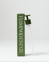 A Flamingo Estate olive-green pump dispenser leans against its branded olive recyclable carton on a soft gray studio background.