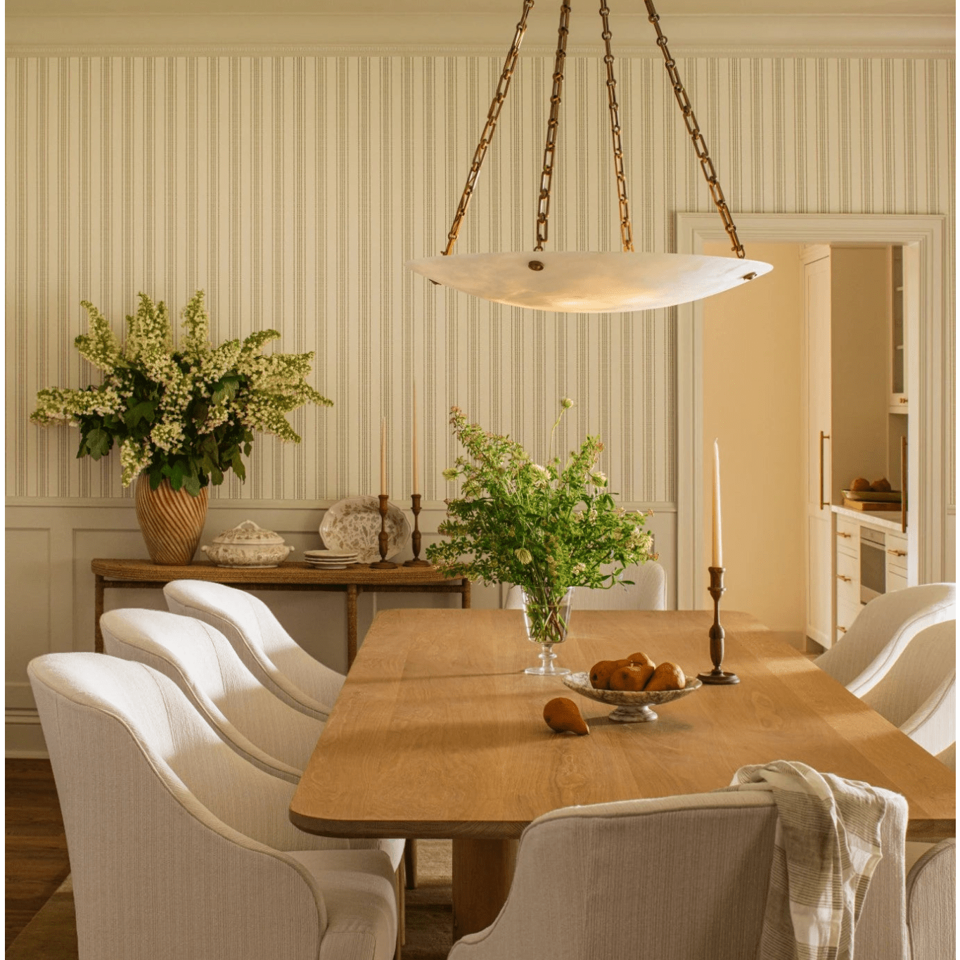 A dining room scene with a light wood rectangular table surrounded by white upholstered chairs, a hanging pendant light, striped wallpaper, and floral arrangements in the background.
