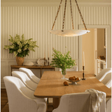 A dining room scene with a light wood rectangular table surrounded by white upholstered chairs, a hanging pendant light, striped wallpaper, and floral arrangements in the background.