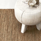Calimia Home River Rug, a handwoven natural jute rug with a rippled texture, placed under a rustic white wood stool.