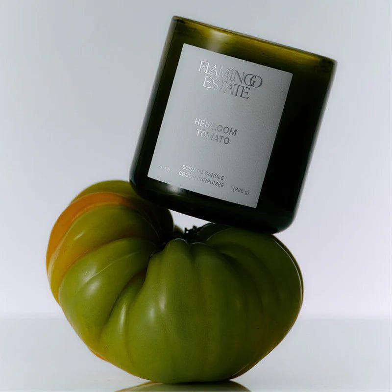 A green glass Roma Heirloom Tomato Candle rests playfully tilted against a large ridged green heirloom tomato, the Flamingo Estate label catching warm light.