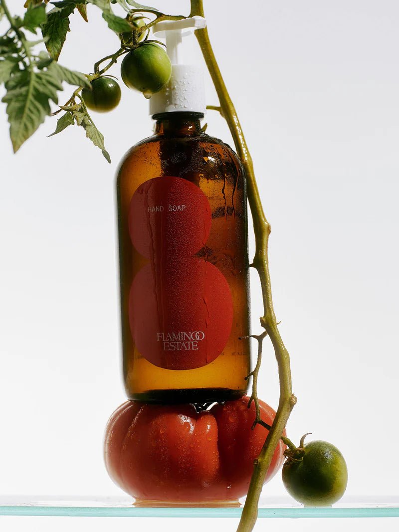 An amber Flamingo Estate Roma Heirloom Tomato Hand Soap bottle perches atop a ripe red heirloom tomato, framed by a fresh green tomato vine.