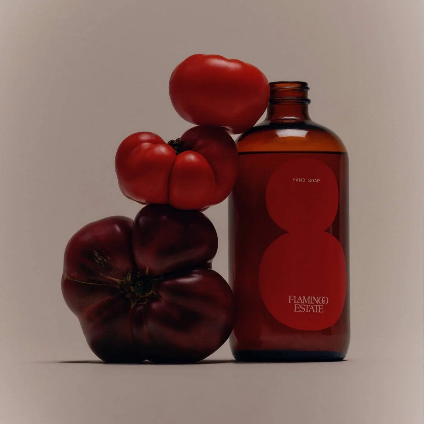 An amber Flamingo Estate Roma Heirloom Tomato Hand Soap bottle stands beside a stack of red and deep purple heirloom tomatoes on a beige backdrop.