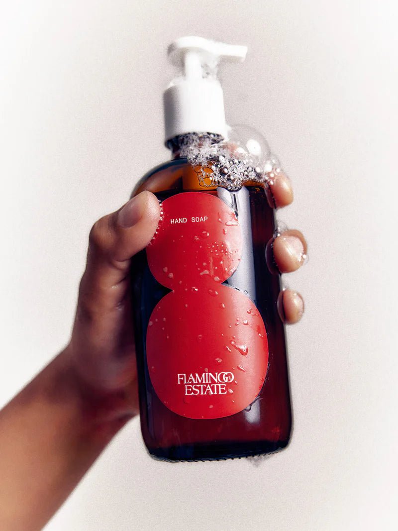 A hand holds the amber Flamingo Estate Roma Heirloom Tomato Hand Soap bottle against a pale wall, glistening with water droplets and a foamy cap.