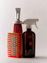A Flamingo Estate Roma Heirloom Tomato Surface Cleaner amber bottle stands beside a vibrant orange and white scrub brush on a bright pale background.