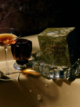 A lathered Flamingo Estate Rosemary & Parsley Soap Brick on a glass soap dish beside a cocktail glass on a bath ledge.