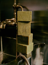 Four green Flamingo Estate Rosemary & Parsley Soap Bricks stacked on a brass bathtub faucet with a shower hose.