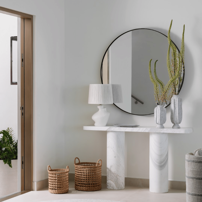 A modern console table with white and gray marble pillars, styled with a white table lamp, two gray textured vases with green stems, and two woven wicker baskets on a light beige floor against a white wall with a large round black-framed mirror.
