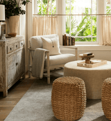 A styled living room corner with a distressed wood cabinet, a cream armchair with a throw pillow and draped blanket, a round beige ottoman table, two woven natural-fiber stools, and a large window with sheer curtains and visible greenery outside.