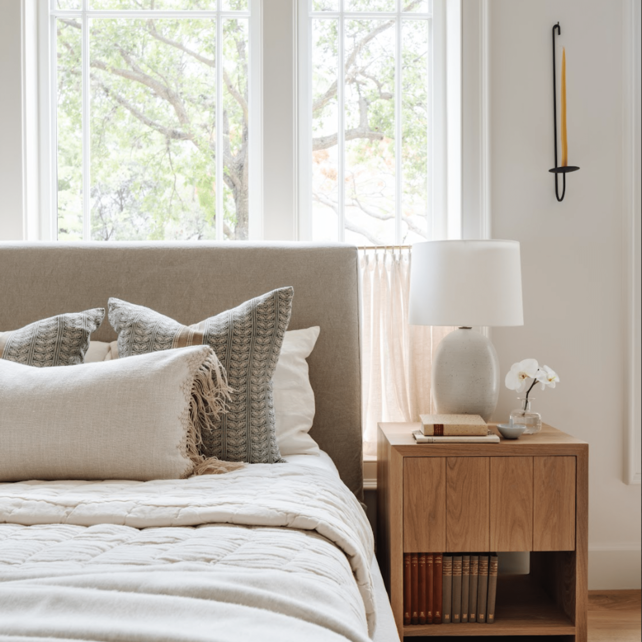 upholstered headboard, layered linen bedding, and patterned pillows. A wooden nightstand holds a ceramic lamp, books, and a small vase with flowers. A single black iron wall candle holder with a beeswax taper hangs beside the window.