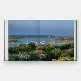 Calimia Home Summer by the Sea book open to a spread showing a harbor full of sailboats beyond coastal gardens.