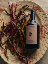 Flamingo Estate Heritage Extra Virgin Olive Oil bottle rests on a fluted ceramic plate piled with long red and green chile peppers.