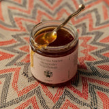 An open jar of Flamingo Estate California Native Mountain Wildflower Honey drips from a gold spoon onto a chevron-woven red and cream mat.