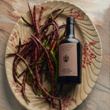 Flamingo Estate Heritage Extra Virgin Olive Oil bottle rests on a fluted ceramic plate piled with long red and green chile peppers.