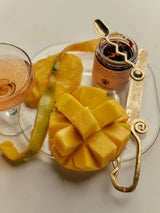 A scored mango and coupe glass share a plate with the Flamingo Estate Salsa Macha Chile Crisp jar and a gold spoon, hinting at sweet-meets-spicy pairings.