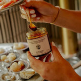 Hands lift a gold spoon from the Flamingo Estate Salsa Macha Chile Crisp jar to drizzle over platters of fresh oysters and shrimp.