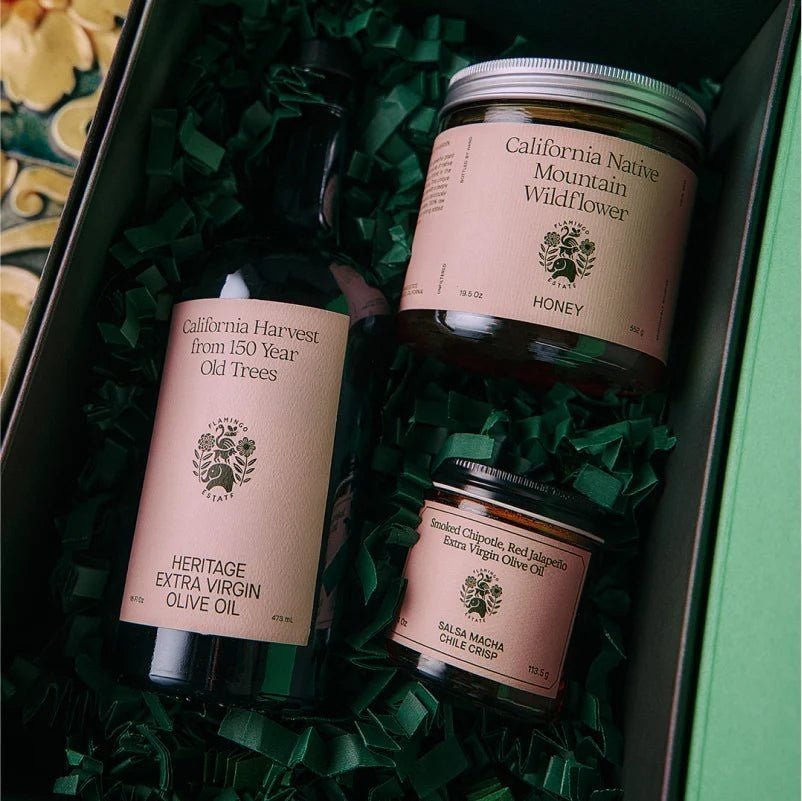 Inside the open Chef's Kiss gift box, the honey jar, olive oil bottle, and salsa macha chile crisp nestle in green crinkle paper from Calimia Home Shop.