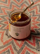 An open jar of Flamingo Estate California Native Mountain Wildflower Honey drips from a gold spoon onto a chevron-woven red and cream mat.
