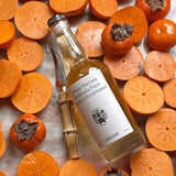 Overhead shot of the Flamingo Estate Persimmon Vinegar laid across whole and sliced Fuyu persimmons with a bone-handled paring knife.