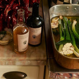 Flamingo Estate Persimmon Vinegar stands beside Heritage olive oil on a vintage marble counter with leeks, garlic, and dried chiles overhead.
